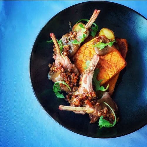 A black plate with lamb chops topped with a savory sauce, served on a bed of polenta and garnished with arugula.
