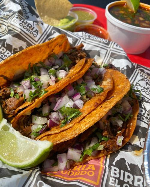 Three tacos filled with meat, topped with onions and cilantro, served with lime and sides of salsa and soup.