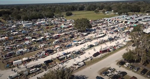 Aerial view of a bustling outdoor market with numerous stalls, vehicles, and visitors in a large open area.