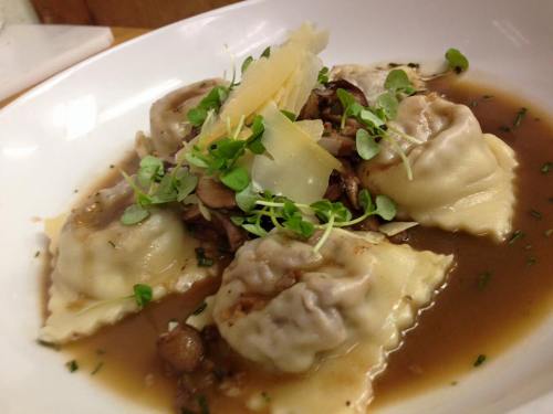 A bowl of ravioli in broth, topped with mushrooms, herbs, and shaved cheese.