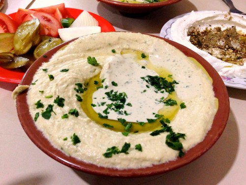 A bowl of hummus drizzled with olive oil and topped with chopped herbs, served with pickles and fresh vegetables.