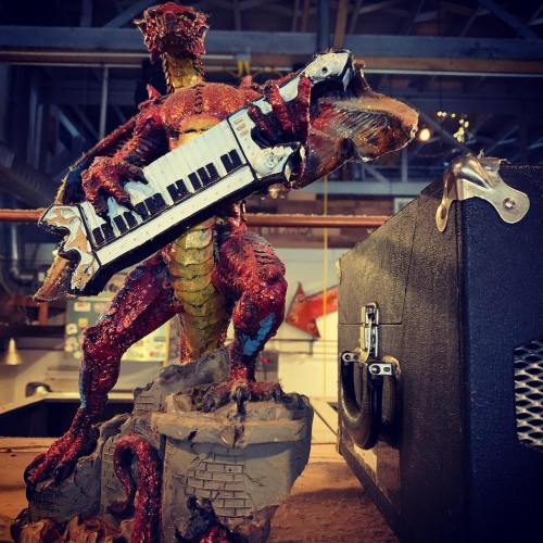 A colorful dragon figurine playing a keytar, set against a rustic background with an amplifier.