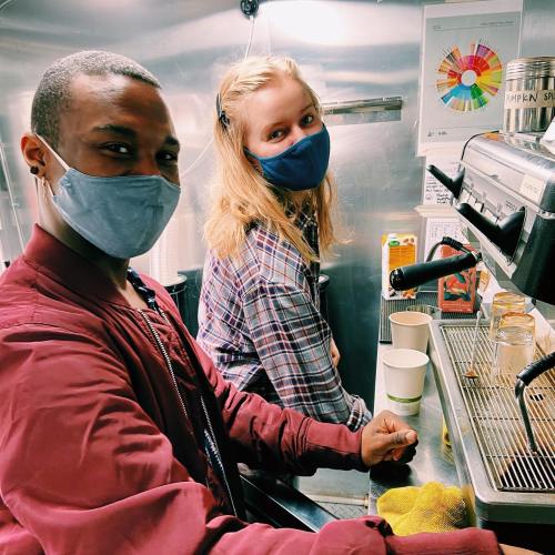 Two people wearing masks smile while working at a coffee machine in a cafe, with cups and supplies around them.