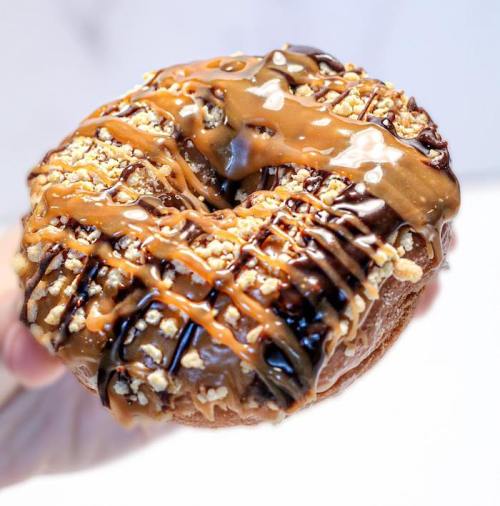 A hand holding a decadent donut topped with chocolate, caramel drizzle, and crushed nuts.