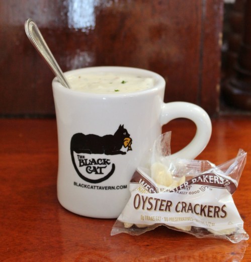 A mug of creamy soup with a spoon, accompanied by a pack of oyster crackers on a wooden table.