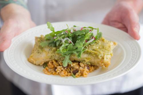 A plate of fish topped with green sauce and arugula, served on a bed of rice with beans.