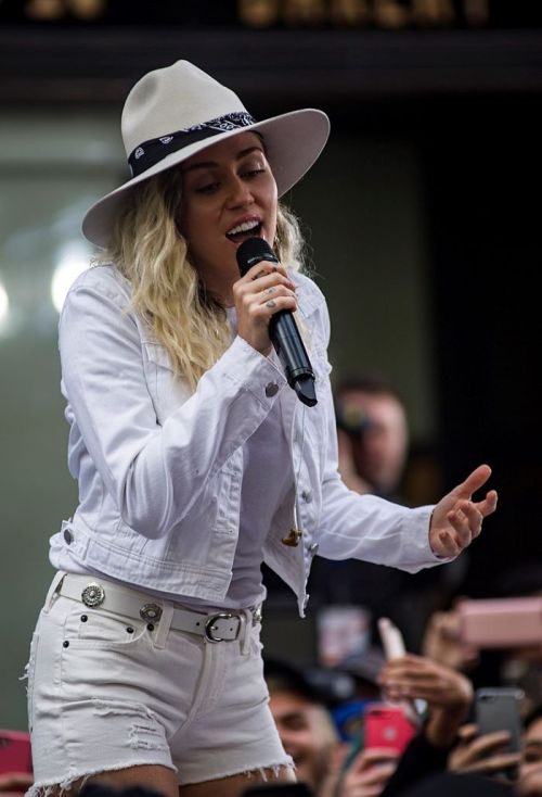 Miley Cyrus performs on stage, wearing a white outfit and hat, singing into a microphone with fans in the background.