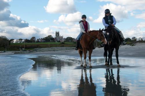 Two riders on horseback stand on a beach, reflecting in the water, with a scenic background of clouds and a distant church.