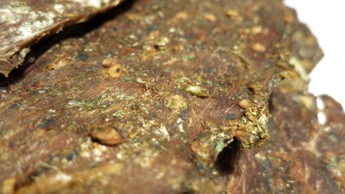 Close-up of textured, brown organic material with small seeds and debris visible on the surface.