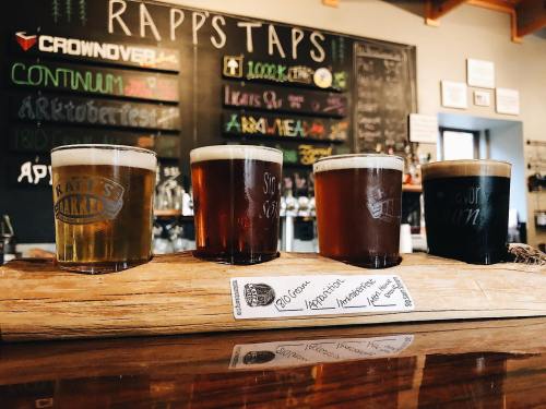 A wooden flight board holds four different craft beers in glasses, set against a bar with a chalkboard menu.