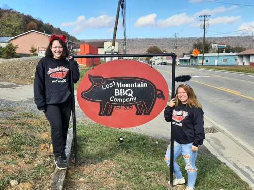 Two women in matching sweatshirts pose beside a sign for Lost Mountain BBQ Company, with a rural backdrop.
