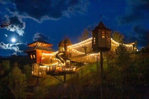 A whimsical treehouse at night, illuminated by string lights, with a slide and surrounded by trees under a full moon.