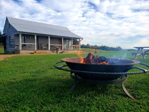 A cozy cabin with a metal roof sits in a grassy field, with a fire pit burning in the foreground under a cloudy sky.