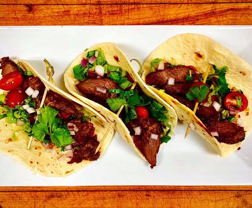Three tacos filled with grilled meat, topped with cilantro, onions, and cherry tomatoes, served on a white plate.