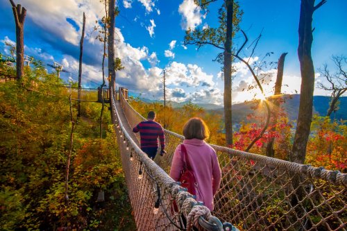 A man and woman walk across a rope bridge surrounded by colorful autumn trees and a scenic sunset.