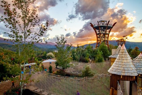 A scenic view of a tower amidst trees, with a sunset sky and playful structures in a mountainous landscape.