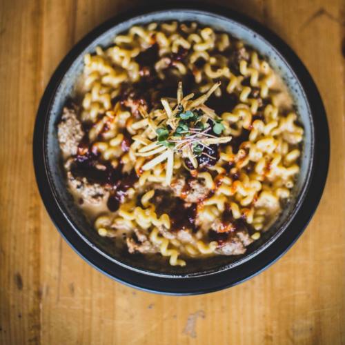 A bowl of pasta topped with meat, sauce, and crispy garnishes, served on a wooden table.