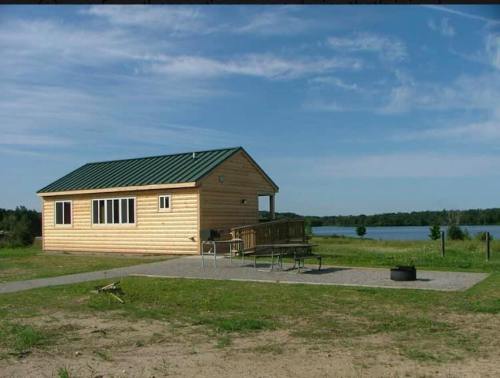 A small wooden cabin with a green roof, surrounded by grass and near a calm lake under a clear blue sky.