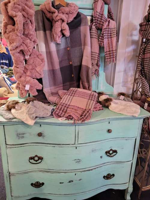 A vintage turquoise dresser displays various cozy scarves and a plaid shirt in a boutique setting.