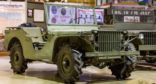 A vintage military jeep with a green exterior, parked indoors among other vehicles and military memorabilia.