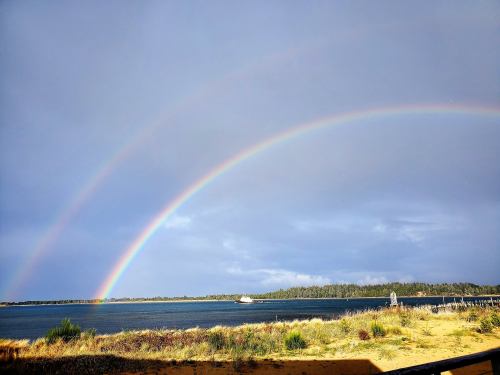 A vibrant double rainbow arcs over a calm lake, with lush greenery in the background under a cloudy sky.