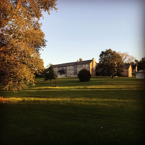 A historic log cabin surrounded by trees and a grassy field, illuminated by warm evening light.