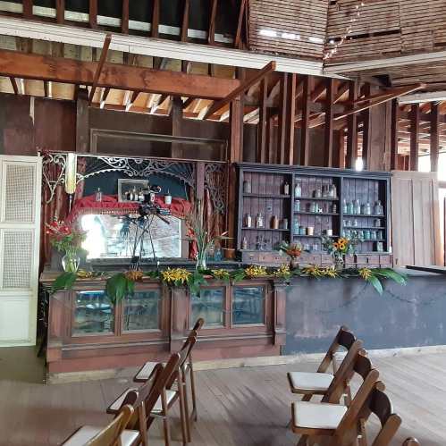 A rustic interior featuring a wooden bar with shelves, plants, and chairs arranged for an event in a vintage setting.