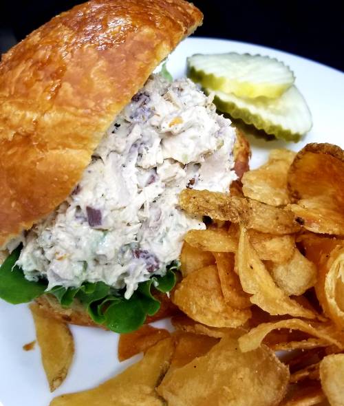 A croissant sandwich filled with chicken salad, served with pickles and a side of crispy potato chips.