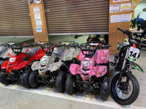 A row of colorful ATVs and a dirt bike displayed in a shop, with a closed garage door in the background.