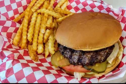 A juicy hamburger with pickles, lettuce, and tomato, served with crinkle-cut fries in a red and white checkered basket.