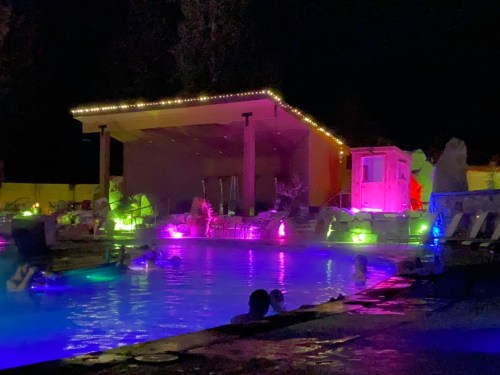 A vibrant pool area at night, illuminated with colorful lights and surrounded by lounge chairs and a modern structure.