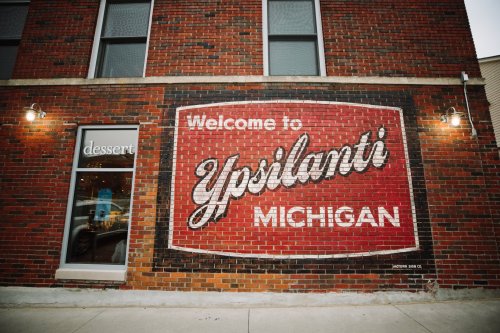 Mural on a brick wall reading "Welcome to Ypsilanti, Michigan" next to a dessert shop entrance.