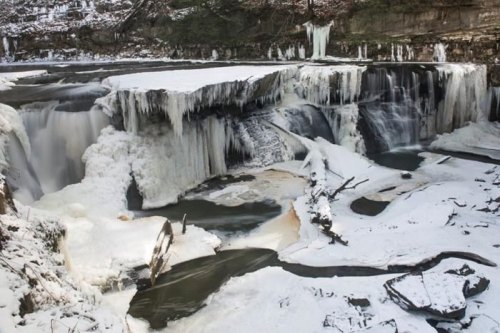 Frozen waterfall surrounded by snow and ice, with rocky cliffs and a partially frozen river below.