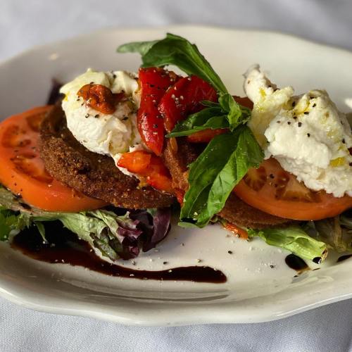 A plate of fried green tomatoes topped with fresh mozzarella, roasted red peppers, basil, and a drizzle of balsamic glaze.