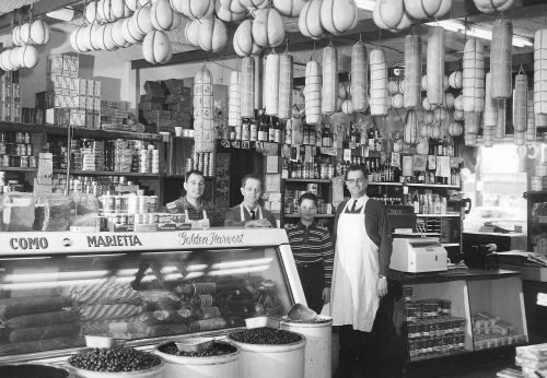 Black and white photo of four men in a vintage grocery store, surrounded by shelves of products and a display case.