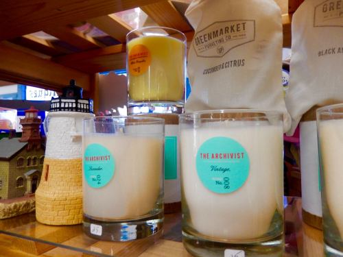 Candles displayed on a wooden shelf, featuring labels like "The Archivist" and "Greenmarket," alongside decorative items.