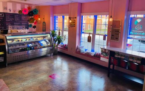 Brightly lit dessert shop with pink walls, a display case of treats, and colorful decor near large windows.