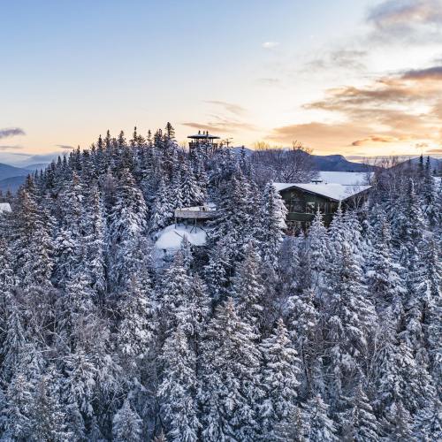 A snowy landscape with evergreen trees and a modern building nestled among them, under a colorful sunset sky.