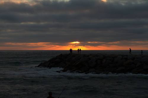 Silhouettes of people on a rocky jetty at sunset, with clouds and waves in the foreground.