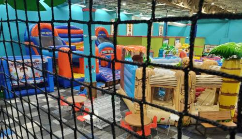 A colorful indoor play area filled with inflatable structures, play equipment, and seating, viewed through a safety net.