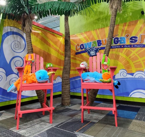 Two colorful chairs with plush toys, surrounded by palm trees and a vibrant backdrop at a playful indoor venue.