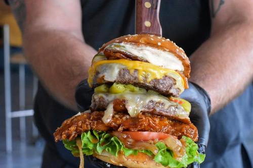 A towering burger with layers of beef patties, fried chicken, cheese, lettuce, tomato, and pickles, held by a gloved hand.