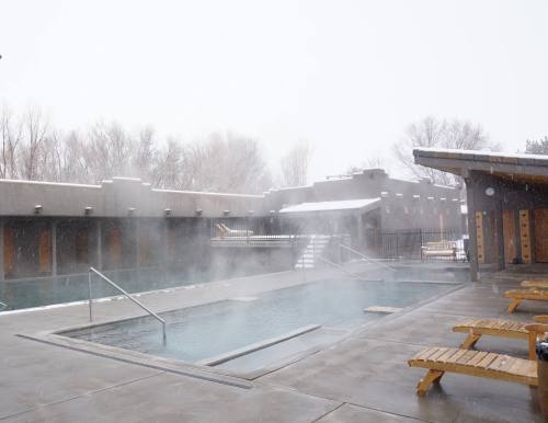 A snowy scene featuring an outdoor hot spring pool with steam rising, surrounded by wooden lounge chairs.