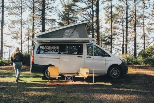 A white camper van with a pop-up roof parked in a forest, with two chairs set up outside.
