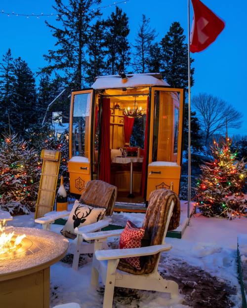 A cozy outdoor scene with a yellow cabin, snow-covered trees, and festive lights, featuring a fire pit and chairs.