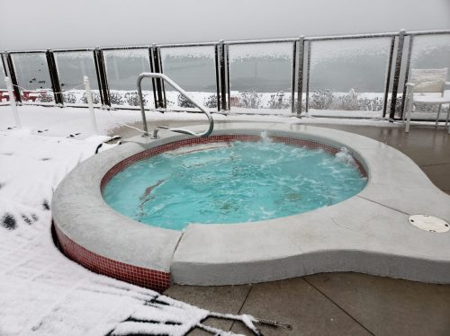 A hot tub with bubbling water sits on a snowy patio, surrounded by a glass railing and a winter landscape.