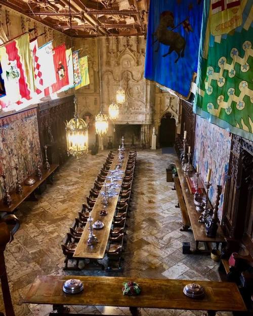 A grand dining hall with long wooden tables, ornate chandeliers, and colorful banners hanging from the ceiling.