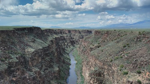 A scenic view of a deep canyon with a winding river, surrounded by green vegetation and distant mountains under a cloudy sky.