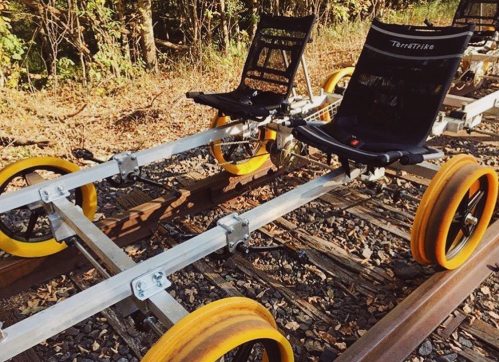 A two-seat rail bike with yellow wheels on a gravel railway track, surrounded by trees.