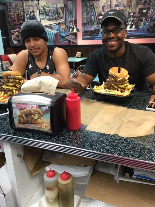 Two men sit at a diner counter with large burgers and fries, smiling and enjoying their meal. Condiments are on the table.